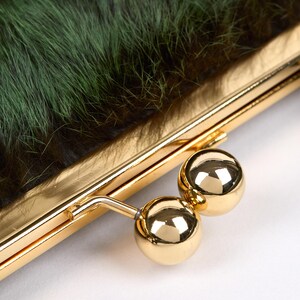 May include: Close-up of a clutch purse with a gold-colored frame and clasp. The purse features a textured, dark green fur exterior. The clasp is a pair of shiny, gold-colored spheres.