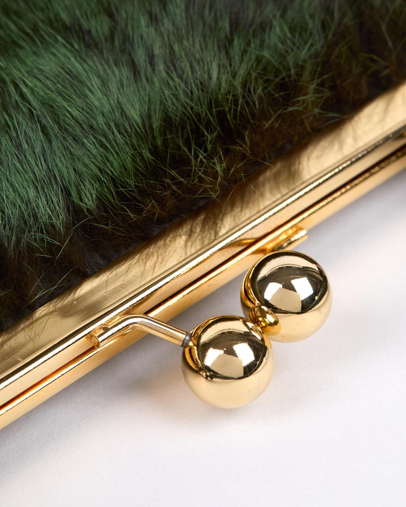May include: Close-up of a clutch purse with a gold-colored frame and clasp. The purse features a textured, dark green fur exterior. The clasp is a pair of shiny, gold-colored spheres.