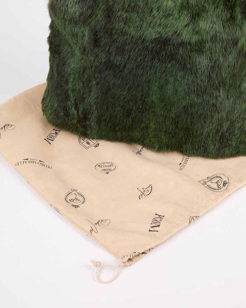 May include: A dark green, textured bag rests on a tan drawstring bag. The dark green bag has a plush, fur-like appearance. The tan bag is printed with black text and logos, and has a drawstring closure.
