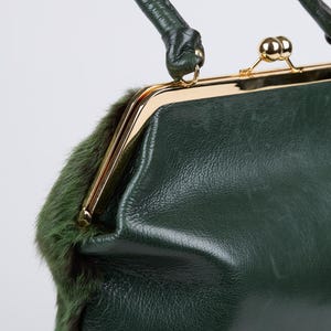 May include: A vintage-style, dark green handbag with a gold-toned clasp and handle. The bag features a textured leather exterior and a strip of green fur trim. The handle is also dark green.