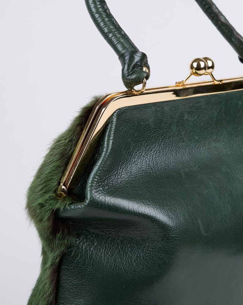 May include: A vintage-style, dark green handbag with a gold-toned clasp and handle. The bag features a textured leather exterior and a strip of green fur trim. The handle is also dark green.