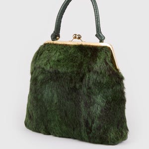 May include: A vintage-style handbag with a dark green fur exterior. The bag features a gold-toned clasp and a matching green leather handle. The bag's shape is rectangular with rounded edges.