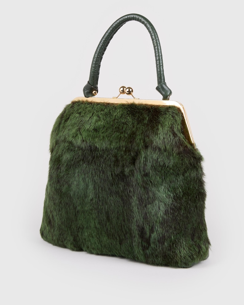 May include: A vintage-style handbag with a dark green fur exterior. The bag features a gold-toned clasp and a matching green leather handle. The bag's shape is rectangular with rounded edges.