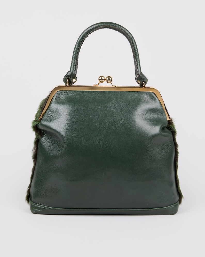 May include: A dark green handbag with a structured frame and a top handle. The bag features a gold-colored clasp and trim, with fur accents on the sides. The bag's design is reminiscent of vintage styles.