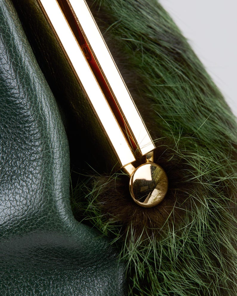May include: Close-up of a green leather handbag with a gold-colored clasp. The bag features a section of green and brown fur. The leather has a textured appearance, and the clasp is a shiny gold color.