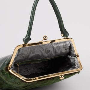 May include: A green handbag with a gold-colored frame and a matching handle. The bag's exterior has a textured, fur-like appearance. The interior is lined with a patterned fabric. The bag is open, revealing the inside.