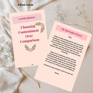 May include: Two open cards with text and floral accents. One card reads "Choosing Contentment Over Comparison" and the other "My Surrender Prayer." The cards are on a light peach background with sprigs of dried flowers.