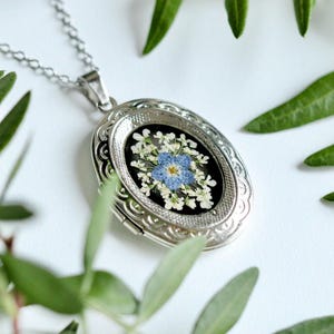 May include: Silver oval locket necklace with a chain. The locket contains a floral arrangement of blue and white dried flowers on a black background. Green leaves surround the necklace.