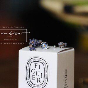 May include: A silver ring with a small, round blue floral design is displayed on a white box. The box has the word "FIGUIER" printed on it. Dried lavender sprigs and the text "aoi haru" are also visible.