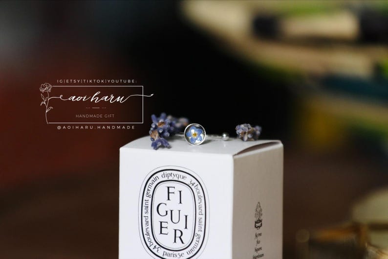 May include: A silver ring with a small, round blue floral design is displayed on a white box. The box has the word "FIGUIER" printed on it. Dried lavender sprigs and the text "aoi haru" are also visible.
