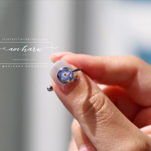 May include: A silver ring featuring a small, round, clear setting with a blue and yellow flower. The ring is held between fingers. The image also includes text that reads "IG: @etsyitiktokyoutube aoi haru HANDMADE GIFT @adiharu handmade".
