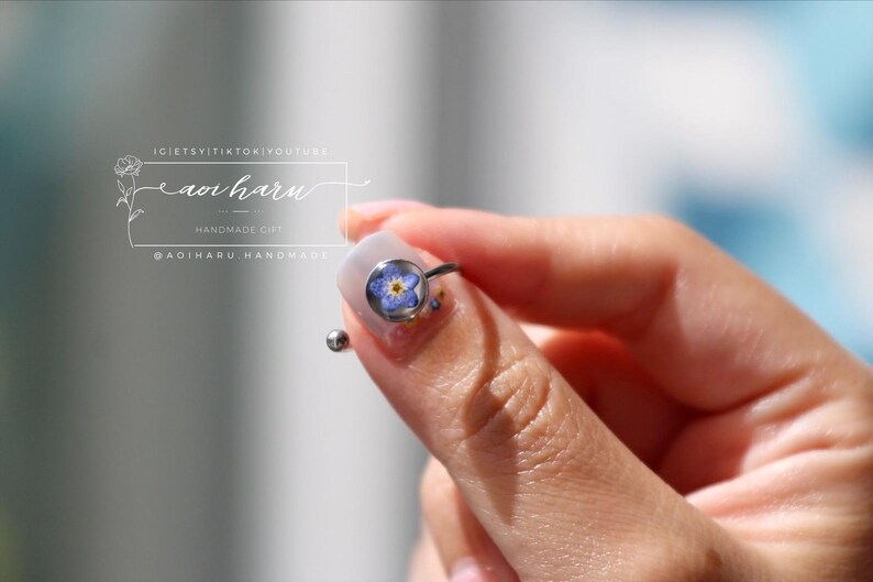 May include: A silver ring featuring a small, round, clear setting with a blue and yellow flower. The ring is held between fingers. The image also includes text that reads "IG: @etsyitiktokyoutube aoi haru HANDMADE GIFT @adiharu handmade".