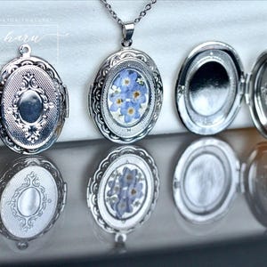 Op de afbeelding: Zilveren ovale medaillons met ingewikkelde details, sommige open om ruimte te bieden voor een foto of klein aandenken. Eén medaillon bevat delicate blauwe bloemen. De medaillons worden gepresenteerd op een reflecterend oppervlak.