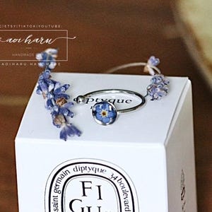 May include: A silver ring with a blue flower set in resin, displayed on a white box. The ring is accented with dried lavender sprigs. The box has the text "diptyque" and "FiGU" printed on it. The image also includes the text "aoi haru".
