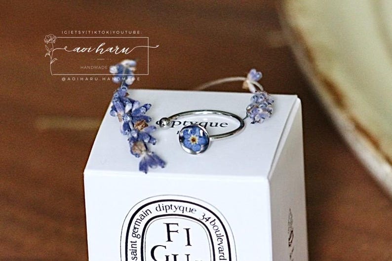 May include: A silver ring with a blue flower set in resin, displayed on a white box. The ring is accented with dried lavender sprigs. The box has the text "diptyque" and "FiGU" printed on it. The image also includes the text "aoi haru".