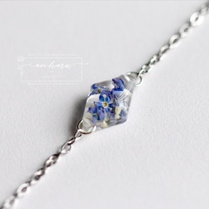 May include: A silver-toned metal chain bracelet featuring a central diamond-shaped clear resin pendant. The pendant contains small, pressed blue forget-me-not flowers with yellow centers. A rectangular logo with the text aoharu is visible.