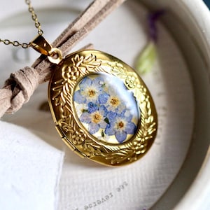 May include: An oval gold-toned metal locket pendant suspended from a delicate gold chain. The locket features an embossed floral border and a clear center display containing pressed blue forget-me-not flowers with yellow centers.