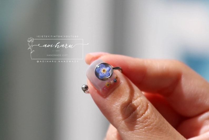 May include: A silver ring featuring a small, round, clear resin setting with a blue and yellow flower. The ring is held between fingers, showcasing the delicate floral design. The ring is a handmade gift.