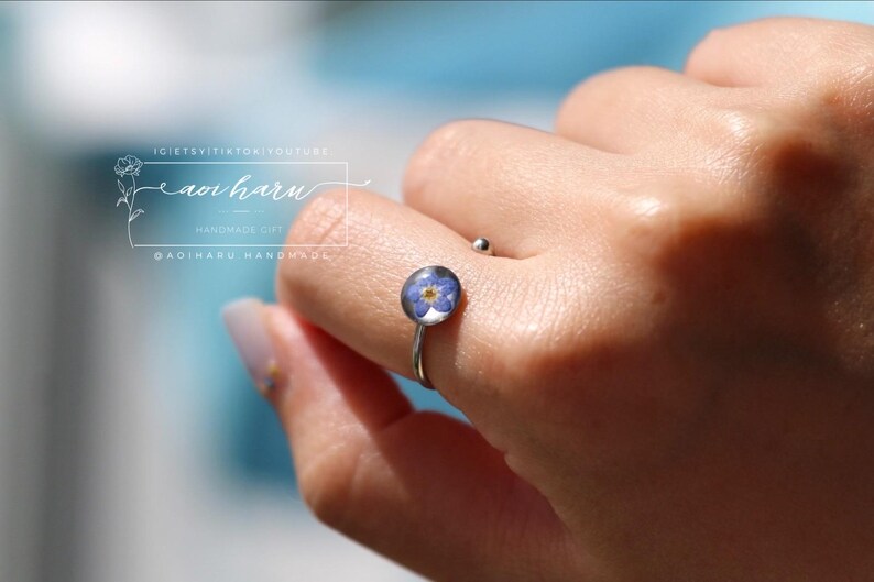 May include: A silver ring featuring a small, round, clear resin setting with a delicate blue and yellow flower. The ring is worn on a finger. The image also includes the text "aoi haru" and other text.