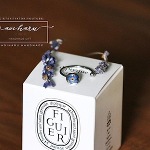 May include: A silver ring with a blue floral design, displayed on a white box with the text "FIGUIER" and other text. The ring is adorned with small purple flowers. The box is on a brown surface.