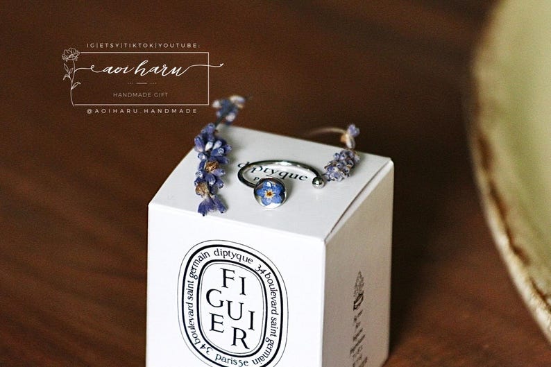 May include: A silver ring with a blue floral design, displayed on a white box with the text "FIGUIER" and other text. The ring is adorned with small purple flowers. The box is on a brown surface.