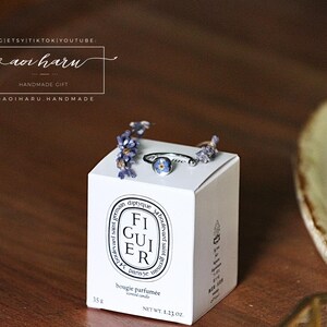 May include: A white Diptyque candle box with the text "FIGUIER" and "bougie parfum&eacute;e" is topped with a silver ring with a blue flower and sprigs of purple flowers. The box is on a wooden surface.