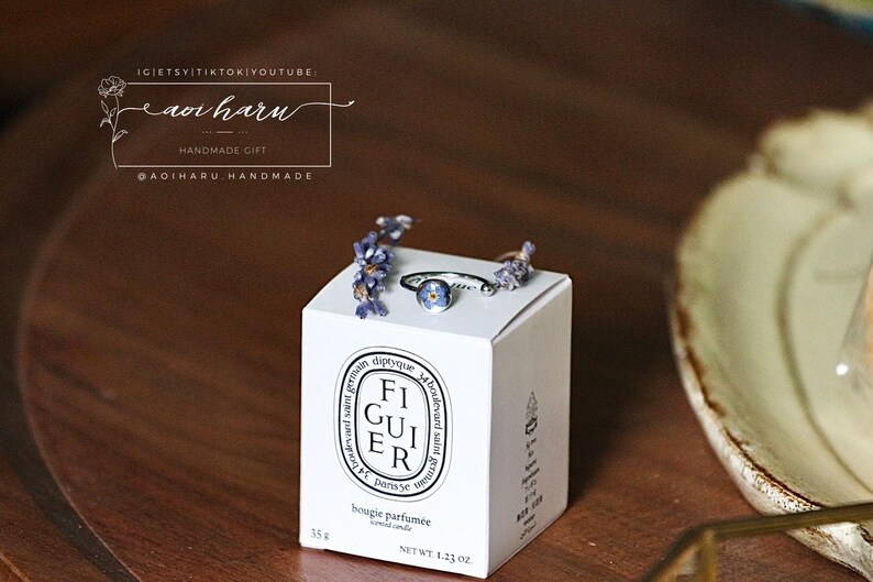 May include: A white Diptyque candle box with the text "FIGUIER" and "bougie parfum&eacute;e" is topped with a silver ring with a blue flower and sprigs of purple flowers. The box is on a wooden surface.