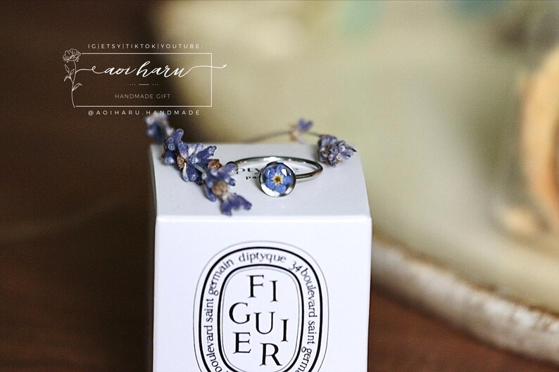 May include: A silver ring with a small, round, blue flower set in clear resin. The ring sits on a white box with the word "FIGUIER" printed on it. Dried lavender sprigs are arranged around the ring. The image also includes text that reads "aoi haru".
