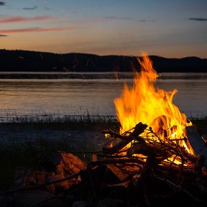 Könnte beinhalten: Ein Lagerfeuer brennt hell mit orangefarbenen und gelben Flammen vor einem dämmerigen Himmel. Das Feuer wird mit Holzscheiten und Ästen gebaut, mit einem See und Bergen im Hintergrund. Die Szene vermittelt Wärme und Ruhe.