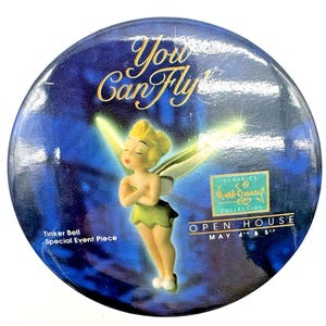May include: A round, blue Tinker Bell collectible with the text "You Can Fly" in gold script. The image features a figurine of Tinker Bell with her arms crossed, wearing a green dress. The text "Classics Disney Collection" and "Open House May 4th & 5th" are also visible.