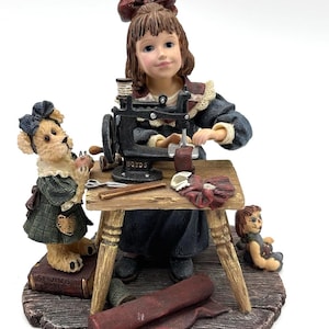 Op de afbeelding: Een beeldje van een jong meisje dat naait aan een houten tafel met een vintage naaimachine. Een teddybeer en een pop zijn in de buurt. Het meisje draagt een donkerblauwe jurk en een rode strik. De naaimachine heeft het woord "BOYDS".