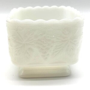 May include: A square white milk glass planter featuring a scalloped top rim and a raised relief pattern of grape clusters and leaves on all sides. The vessel sits on a square pedestal base and has a textured, lace-like background detail.
