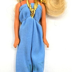 May include: A vintage doll with blonde hair and a blue jumpsuit. The doll has a gold necklace and matching hair accessory. The doll is wearing blue heels. The doll is set against a white background.