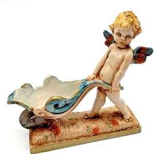 Puede incluir: Figura de querubín empujando una carretilla decorativa. La figura tiene cabello rubio y alas azules y rojas. La carretilla es blanca con detalles azules y dorados, y descansa sobre una base rectangular con detalles florales.