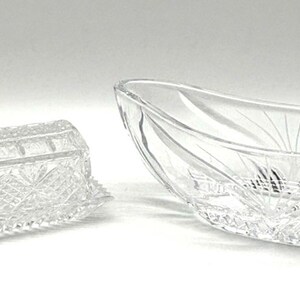 May include: A clear, rectangular glass butter dish with a lid and a decorative, starburst pattern. Beside it is a clear, oval-shaped glass serving bowl with etched leaf and sunburst designs. Both items are on a white surface.