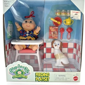 Può includere: Un set da gioco per l'alimentazione da collezione Cabbage Patch Kids. Il set include una bambola su un seggiolone, un cane giocattolo e vari accessori per l'alimentazione. La confezione presenta un motivo a scacchi rossi e bianchi e la scritta "Feeding Playset".