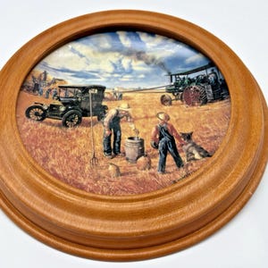 Puede incluir: Plato decorativo redondo con marco de madera. El plato presenta una escena campestre con un coche antiguo, un tractor y personas trabajando en un campo. La obra de arte utiliza tonos cálidos de marrón, amarillo y azul.