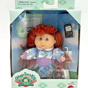 May include: A Cabbage Patch Kids collectible doll in its original packaging. The doll has red yarn hair, blue eyes, and wears a purple and floral dress. The box includes accessories and the text "Collectible" and "For ages over 3 years."