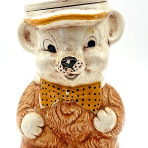 May include: A vintage ceramic bear cookie jar. The bear is tan with a white face, wearing a yellow bow tie with black polka dots and a white hat with a yellow brim. The lid has a wooden knob.