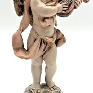 Puede incluir: Figurilla detallada de un querubín tocando una lira. El querubín está representado con alas y envuelto en una tela fluida. La figurilla está de pie sobre una base decorativa, con una paleta de colores beige, marrón y toques de rosa.