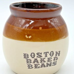 May include: A small, ceramic Boston Baked Beans crock. The jar has a cream-coloured base with a brown band and a dark brown rim. The words "BOSTON BAKED BEANS" are printed in a dark grey, vintage font.