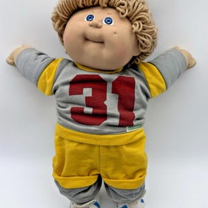 Op de afbeelding: Een vintage Cabbage Patch Kid pop met krullend blond haar en blauwe ogen. De pop draagt een grijs en geel voetbalshirt met het nummer 31 in rood en een bijpassende korte broek. Witte sneakers met blauwe strepen maken de outfit compleet.