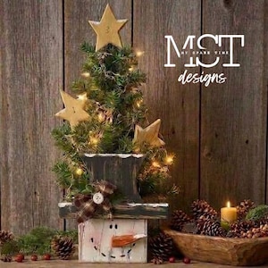 Könnte beinhalten: Ein künstlicher Tisch-Tannenbaum, geschmückt mit warmweißer Lichterkette und drei Holzsternen. Der Baum steht in einem quadratischen Sockel, der wie ein Schneemanngesicht bemalt ist und eine karierte Stoffschleife trägt. Der Text MST My Spare Time designs ist sichtbar.