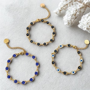 May include: Three gold-toned chain bracelets with an evil eye design. Each bracelet features a series of small, circular charms with blue and black eye details. The bracelets are displayed on a white surface.