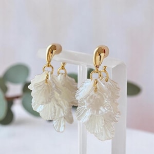 Può includere: Un paio di orecchini eleganti con borchie color oro e pendenti bianchi a forma di petalo. Gli orecchini sono esposti su un supporto trasparente, con uno sfondo morbido e sfocato.