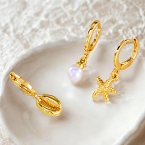 May include: Gold-colored hoop earrings with ocean-themed charms. One earring features a pearl, another a starfish, and the third a seashell. The earrings are displayed on a white, textured surface.
