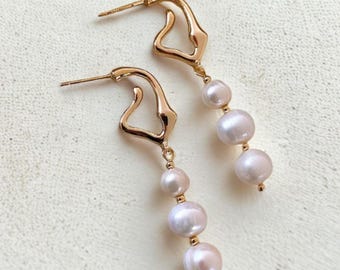 May include: A pair of gold-tone earrings with an abstract design. Each earring has a curved, open shape at the top, with three light pink pearls hanging below. The pearls vary in size, creating an elegant, flowing appearance. The earrings are suitable for various occasions.