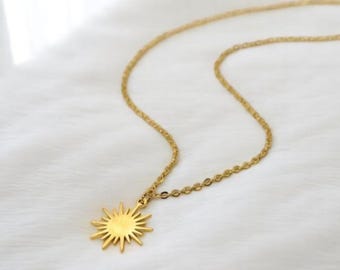May include: A gold-colored necklace with a sunburst pendant. The pendant has a detailed sun design with rays extending outwards. The chain is delicate and appears to be made of small, linked segments. The necklace is displayed on a white, textured surface.
