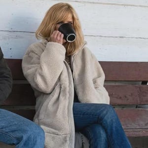 May include: A long, cream-colored, zip-up fleece jacket with a textured, fuzzy exterior. The jacket features a high collar, long sleeves, and a straight hemline. It is worn over blue denim jeans, paired with brown boots, while holding a black mug.
