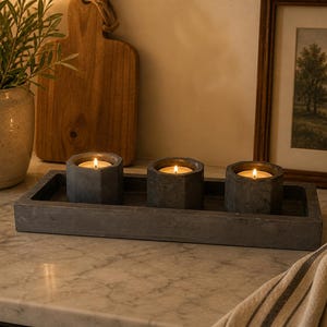 May include: Three lit gray hexagonal candles sit in a matching rectangular tray. The candles are on a marble countertop, with a framed landscape painting and a wooden cutting board in the background. A potted plant is to the left.